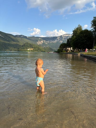 The media file shows a toddler standing in shallow, clear water with a mountain range and a lake in the background. The child, facing away from the camera, has blonde hair and is wearing a swim diaper. The water is rippled, reflecting the sunlight, and the rocky lakebed is visible beneath the surface.
The scene is set in Novalaise, France, as suggested by the location context. The background features a lush green mountainside dotted with trees, leading up to rocky peaks. A bright blue sky with scattered white clouds suggests it is daytime, likely late morning or early afternoon, given the quality of light. The presence of boats on the lake and people on the shore indicates a recreational area.
The overall mood is peaceful and idyllic, evoking a sense of summer leisure and childhood joy. The focus on the child's interaction with the water, combined with the scenic natural backdrop, creates a serene and pleasant atmosphere. There is no visible text in the media file.