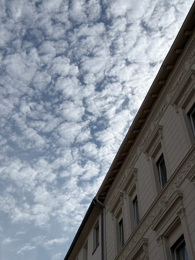 This image shows a section of a cream-colored building with ornate architectural details, including decorative moldings and cornices. The building is viewed from a low angle, with its roofline and upper floors visible. The sky above is filled with scattered white clouds against a light blue background. The image appears to be taken in Witten, Germany, based on the provided context, though no specific landmarks are identifiable within the frame. There are no people or vehicles visible in the scene.