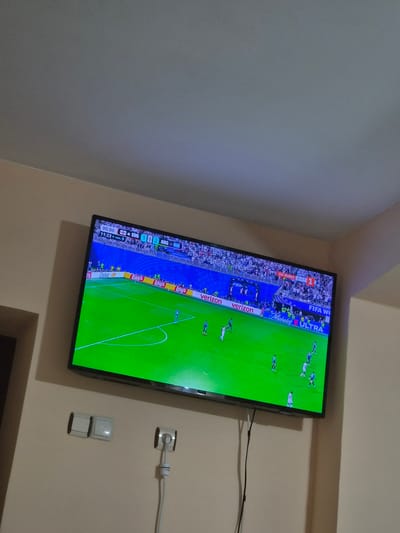 A television screen displays a soccer match, with the score showing 1-1 between ENG and ARG in the 90th minute, plus 9 minutes of added time. The field is illuminated by bright green grass. Several players in blue and white uniforms are visible on the field. The screen also shows advertising for brands like Dove, Lay's, and Verizon. The television is mounted on a light-colored wall. Below the television are two light switches and an electrical outlet with a white cord plugged in. The ceiling above is a neutral gray.