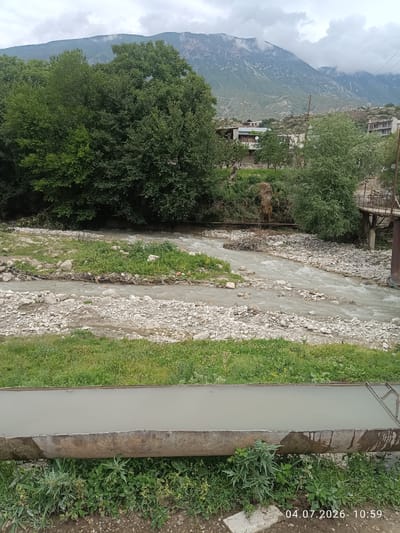 In the city of Rakhata, Russia, a cloudy day frames a picturesque landscape dominated by a river flowing through a rocky, vegetated area. Majestic mountains loom in the background, partially obscured by mist, with a small village nestled in the valley below. The foreground features a man-made trough filled with water, surrounded by lush green grass and various plants. A bridge crosses the river in the mid-ground, hinting at the human presence in this natural setting. The scene is captured on July 4, 2026, at 10:59 AM.