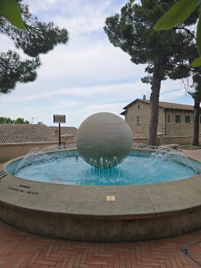 In the city of San Marino, San Marino, a large, spherical fountain centerpiece is prominently displayed within a circular fountain basin. The sphere, made of a textured, light-colored material, sits at the center of the basin, which is filled with bright blue water. Water streams upward from multiple spouts around the sphere, creating a dynamic visual effect. The fountain is constructed from a pale, concrete-like material, with visible inscriptions along its rim. One inscription reads "INAUGURATA IL 21 MAGGIO 1962 - 1661 D.F.R.", indicating its inauguration date. Another small plaque, mounted on the fountain's edge, states "ACQUA NON POTABILE - NON-POTABLE WATER" in both Italian and English. The fountain is situated on a patterned brick walkway. In the background, traditional buildings with terracotta tile roofs are visible, suggesting a historic urban setting. Evergreen trees with dark green foliage are present in the foreground and frame the scene. The sky above is partly cloudy, with white clouds scattered across a pale blue expanse.