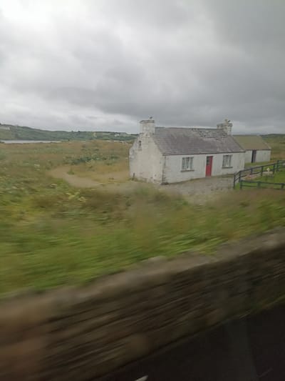 The image depicts a rural landscape under a cloudy sky, featuring a white cottage with a red door. The cottage has two chimneys and appears to be connected to a smaller, adjacent structure with a green roof. A dark wooden fence encloses a grassy area where a white sheep is visible. The foreground shows a blurred view of a stone wall and vegetation, indicating motion. The scene is set in Mullaghduff Scotch, Ireland.