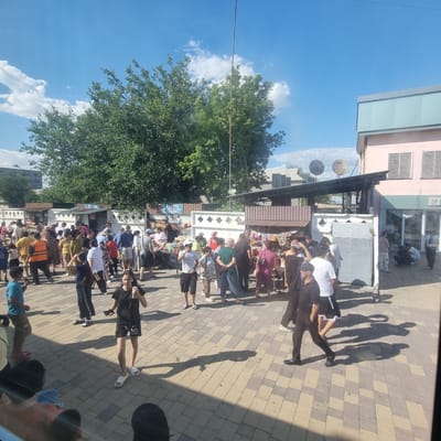 The image shows a bustling open-air market scene in Turkistan, Kazakhstan, on a bright, sunny day with a blue sky and scattered white clouds. A large, leafy green tree dominates the middle ground, providing shade over a paved area where numerous people are gathered. Several market stalls with awnings are set up, displaying a variety of goods, including food items and packaged products. People of various ages and genders, dressed in casual clothing, are browsing, interacting, and walking through the market. Some individuals are wearing traditional headwear. In the background, there are buildings with distinct architectural features, including a pink building with tinted windows and an upper floor with a pale green facade. Satellite dishes are visible on one of the buildings. The ground is paved with interlocking square tiles in beige and grey tones, casting long shadows from the sun. The overall impression is one of a lively community gathering and commercial activity.