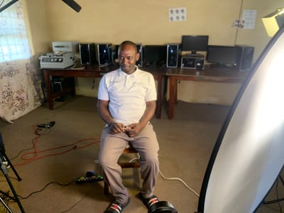 The image shows an indoor scene, likely a room in Gindiri, Nigeria, serving as a makeshift office or tech hub. A man in his late 30s to early 40s, wearing a light-colored polo shirt and khaki pants, is seated on a wooden chair. He has short, dark hair and is looking slightly down and to his left. His hands are clasped in his lap.
The room is cluttered with electronic equipment. On wooden tables in the background, there are multiple computer towers and monitors, suggesting a setup for a computer lab or a video editing station. To the left, near a window with floral curtains, sits a piece of equipment that appears to be an industrial machine or a projector, possibly part of a technical setup.
On the floor, in the foreground and extending towards the seated man, are various cables and electrical accessories. Red and black cords snake across the carpeted floor, connecting to a black power strip with illuminated buttons and switches. A microphone is positioned on the floor to the left, connected by a cable. A camera tripod is also visible on the left side of the frame.
A large, bright white softbox, likely part of a lighting setup for filming or photography, dominates the right side of the image, casting a strong light into the scene. The walls of the room are painted a pale yellow. Small, framed pictures or posters are affixed to the wall in the background. The overall impression is of a functional, albeit somewhat disorganized, technological workspace.
