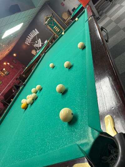 The image shows a pool table with billiard balls on a green felt surface. The table is surrounded by the interior of a establishment, visible through the skewed perspective, that features a sign with the word "PIRAMIDA" in Cyrillic alphabet. The lighting appears dim, and the decor includes visible wall decorations, possibly frames or pictures. The floor and ceiling are visible, suggesting the camera position is elevated and angled downwards at the table. I don't have information about location in the city of San Vicente Del Raspeig/sant Vicent Del Raspeig, Spain.