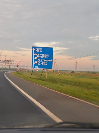 The image shows a roadside view with a blue directional sign on the right. The sign displays text in Russian and English, indicating directions to PSKOV, KUROVITSY, and GATCHINA. Below "GATCHINA", it says "300m". The sign has arrows indicating different routes. The environment includes a paved road with a white line on the left, a grassy shoulder, and distant power lines and transmission towers under a cloudy sky at dusk. The location is identified as Torfyanoye, Russia.