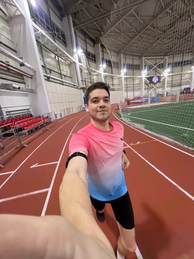 The image shows a man taking a selfie indoors at what appears to be an athletic facility, specifically a running track. The photograph is oriented with the man lying on his back, looking up, and his body positioned diagonally across the frame, suggesting it was taken from a selfie stick or outstretched arm held at an angle and then rotated, or perhaps a camera mounted on his body.
The man, who appears to be in his 20s or 30s, is smiling faintly and looking directly at the camera. He has short, dark hair and is wearing a long-sleeved athletic shirt that features an ombre design, transitioning from pink at the top to light blue at the bottom. He is also wearing dark, possibly black, athletic shorts or leggings. A dark-colored smartwatch or fitness tracker is visible on his left wrist, and a white earbud is in his right ear. His right arm, which is holding the camera, is partially visible in the bottom right corner of the frame.
The background reveals a large indoor sports arena. The dominant feature is the reddish-brown running track with white lane markings. To the left of the man (or above him in the rotated image), there's a green artificial turf field, partially visible, with white netting separating it from the track area. A white soccer goal is also visible on the field.
Further in the background, a large, intricate ceiling structure made of metal trusses and supports is visible, typical of large indoor sports complexes. Numerous bright lights illuminate the facility. On the far side of the track, there are tiered red seats, indicating spectator stands. High above the track and field, a digital display or scoreboard is visible, showing a circular graphic with multiple colors, though specific details or text are indistinguishable. The overall impression is a modern, well-lit indoor sports complex where the man is likely engaged in or preparing for physical activity.