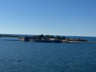 The image displays a wide-angle, eye-level view of the island fortress of Kungsholmen in Karlskrona, Sweden, on a clear, sunny day. The fortress, characterized by its robust stone walls and buildings, is situated on a strip of land extending into the deep blue sea. The water is calm, with subtle ripples reflecting the bright sky. Lush green trees and vegetation cover parts of the island, interspersed with structures of the fortress, including what appears to be a central tower. A long, stone-lined pier juts out from the island, with a small yellow buoy visible at its end. In the distance, the horizon is a clear, unbroken line between the sea and the pale blue sky. There are no people or animals visible in the scene. The overall impression is one of historical significance and natural beauty, with the fortress standing as a prominent landmark against the serene maritime landscape.