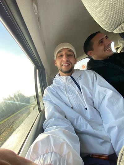 Two adult males are observed inside a moving vehicle, likely a passenger van or car, in La Wantzenau, France, during daylight hours under a clear sky. The primary subject, positioned in the foreground and facing the camera, is a male with a short dark beard and dark hair, wearing a light beige knit beanie and a white hooded anorak jacket. He is smiling broadly, exposing his teeth, and appears to be taking a selfie. His left arm is visible in the lower left corner. Behind him, to the right, a second male is partially visible. This individual also has short dark hair and is smiling while looking towards the right side of the vehicle, away from the camera. He is wearing a dark-colored jacket.
The interior of the vehicle features a light-colored headliner. A large window on the left side of the vehicle reveals a bright, sunlit exterior. The landscape outside consists of a field, possibly tilled, next to what appears to be a road or path. Beyond the field, a line of deciduous trees or bushes with bare branches is visible against the bright sky. The blurred appearance of the exterior suggests the vehicle is in motion. A white circular sticker with a red 'A' is affixed to the window between the two individuals, commonly indicating a novice driver in France. A small, indistinct brown object is visible on the foreground subject's lap.