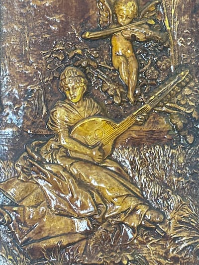 This is a close-up, highly textured image of a relief carving. The carving depicts a scene with multiple figures, rendered in a golden-brown hue with significant depth and shadowing, suggesting it is a bas-relief or high-relief sculpture.
In the foreground, a reclining nude figure is visible, with its body positioned horizontally across the frame. To the left of this figure, another nude figure is depicted, lying on its side. A musical instrument, possibly a lute or lyre, is held by a figure that is partially obscured. This figure appears to be seated or leaning against something, with drapery surrounding it.
Further to the right, another figure is visible, draped in fabric that creates a sense of movement and volume. This figure's face is partially visible, looking upwards. The background of the carving is intricate, featuring what appears to be foliage, branches, and possibly other figures or objects, all rendered with a rough, organic texture.
The overall scene evokes a sense of classical mythology or allegory, with nude figures, musical instruments, and drapery. The lighting accentuates the relief, creating strong highlights and deep shadows that give the carving a sense of drama and three-dimensionality. The material appears to be metal or a similarly hard, moldable substance. There is no discernible text within the image. The context of Пярэжырскі сельскі Савет, Belarus, does not provide any immediate clues to the specific subject matter of this carving.