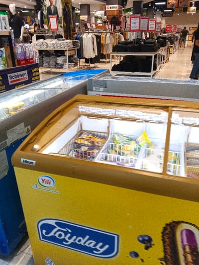 This image captures an indoor scene within a large retail store, likely a hypermarket or department store, located in Sindanglaya, Indonesia. The foreground is dominated by several commercial chest freezers. A prominent yellow freezer, branded "Yili" and "Joyday" with the "Hiron" logo, displays various packaged frozen treats and ice cream bars through its clear glass lid. To its left, a blue freezer, partially visible, carries the "Liebherr" brand. Another silver-colored freezer is also in view.
In the midground and background, the store transitions to a clothing section. Numerous racks are filled with folded and hanging apparel, including t-shirts and shirts, arranged for display. Mannequins, such as one on the far left, showcase some of the clothing items. Promotional signage is abundant; a blue banner for "Robinson mart" is visible, along with several red signs advertising "HARGA LEBIH MURAH!!" (cheaper price) and discounts, including one for "30%" and prices like "245" and "275." Other signs with text like "S135" and "75" are seen in the distance.
A few blurry figures of shoppers or store staff can be observed moving among the clothing racks in the background. The store is brightly illuminated by artificial lighting. The combination of local retail chain names like "Robinson mart" and Indonesian text confirms the geographic context.