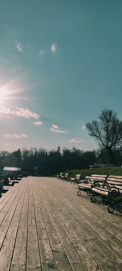 The image shows an outdoor scene in Grebnevo, Russia. A weathered wooden walkway extends into the distance, lined with park benches. The sky is light blue with sparse white clouds, and the sun is shining brightly. Trees and greenery border the walkway.