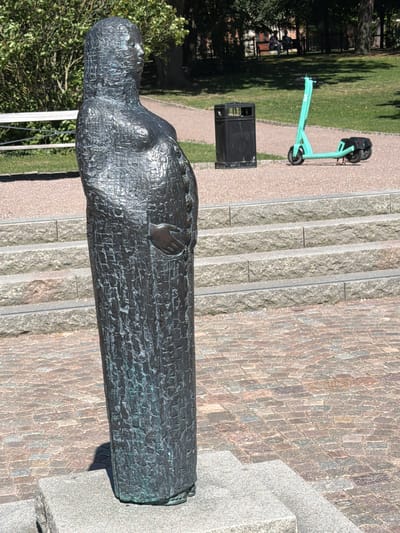 In Lund, Sweden, a weathered bronze statue of a pregnant woman stands on a stone pedestal in a park. The statue is depicted in a long, textured robe, with one hand resting on her abdomen. Behind the statue, a black trash can sits next to a bright green electric scooter. In the background, there are stone steps leading up to a grassy area with trees and benches where two people are seated. The ground is paved with cobblestones.