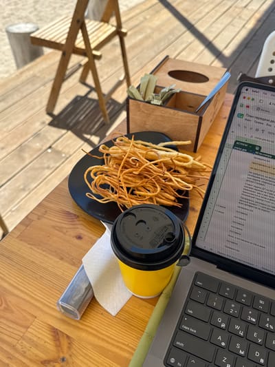 A close-up, eye-level shot depicts a wooden table outdoors, likely at a cafe or restaurant, with a view of a body of water in the background, suggesting the location is Koshkol', Kyrgyzstan. In the foreground, a laptop is open, displaying a spreadsheet with Russian text. Next to the laptop is a yellow disposable coffee cup with a black lid. To the left of the cup, a black plate holds a pile of fried string cheese sticks. Behind the plate and cup is a wooden box containing napkins and sugar packets. A folded wooden chair is visible in the background, casting a shadow on the wooden deck. The overall scene suggests a casual work or break session in an outdoor setting.