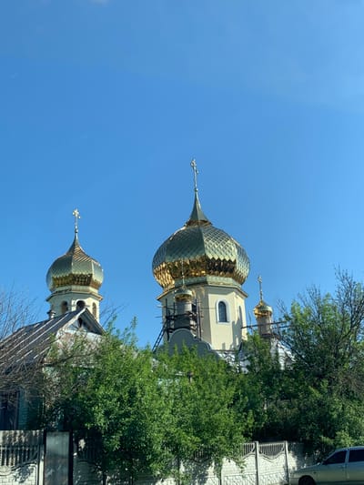 The image shows the front of a church with three gold domes in Kolomak, Ukraine, located at the coordinates 49.837 latitude and 35.294 longitude. The church is made of yellow brick with white trim and has a scaffolding structure on its front. It is partially obscured by a thick line of deciduous trees with green leaves. There is a low brick wall with a metal fence in front of the church, and the top of a car is visible in the bottom right corner of the image. The sky is a bright blue with a few scattered white clouds. The only human subjects are those who built the church and scaffolding, who are not visible in the image.