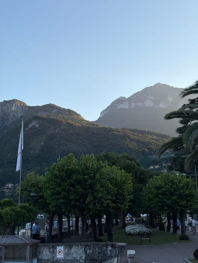 The image depicts a scene in the city of Loveno, Italy, with a prominent view of mountains and a clear sky. In the foreground, several trees with lush green foliage line a pathway. To the left, a white flag flies on a flagpole. In the middle ground, there are indications of a park area with flower beds and more trees. Several people are visible walking and interacting in the area, dressed in casual summer attire. The background is dominated by large, verdant mountains, with some houses nestled on their slopes. The overall atmosphere suggests a pleasant, possibly late afternoon or early evening setting.