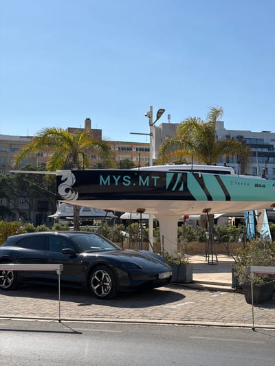 The image depicts a sunny day in Floriana, Malta, with a view of a dark-colored electric vehicle parked on a paved surface. In the background, a modern-looking sailboat with the prominent text "MYS.MT" and "TAUSS B&G" on its hull is on display, supported by structures. Palm trees and buildings are visible behind the sailboat, suggesting an urban or waterfront setting. The vehicle's license plate reads "GNP 710". There are no people visible in the image. The overall scene suggests a commercial or promotional event, possibly related to sailing or automotive industries.