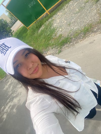 A young woman with long, dark hair, wearing a white baseball cap with a blue emblem and a white, textured button-down shirt, smiles as she takes a selfie outdoors. She is standing on a paved surface, with a gravelly area and green grass to her left, and a green fence with yellow trim behind her. The overall scene suggests a casual, sunny day in Akyar, Russia.