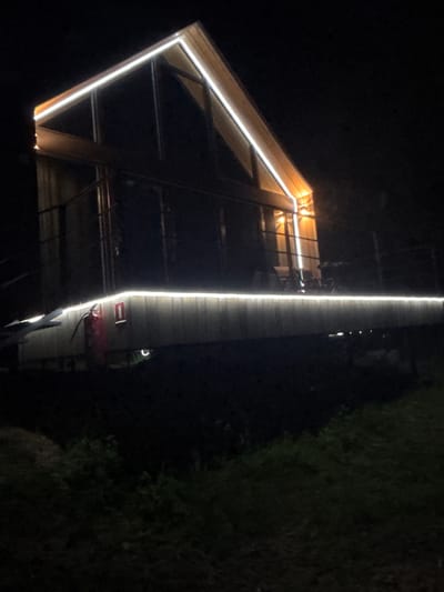 A modern wooden cabin, illuminated by white and orange LED strip lights, is visible at night. The cabin features a steeply pitched roof with white lights outlining its edges. A deck, also lined with white lights, is situated in front of the cabin. On the deck, a red fire extinguisher is mounted on the wooden wall. The surrounding environment appears to be a dark, grassy area, suggesting a rural or semi-rural setting, potentially on the outskirts of Mishkino, Russia. The image is dimly lit, with the cabin and its lighting being the primary focal points.