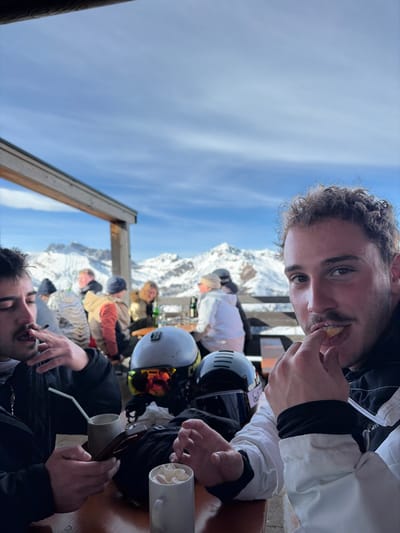 The scene depicts an outdoor dining area at a ski resort in Tux, Austria, under a clear, sunny winter sky. Snow-capped mountains are visible in the background.

In the foreground, two males are seated at a wooden table. The male on the left, with dark hair and a mustache, is actively smoking a lit cigarette held in his right hand, while his left hand holds a dark-colored smartphone. A light-colored ceramic mug with a straw is on the table in front of him. The male on the right, possessing light-brown curly hair and a mustache, looks directly towards the viewer. He holds a small, light-colored piece of food between the fingers of his right hand, bringing it towards his mouth. He is dressed in a white and black ski jacket.

On the table between the two individuals are two ski helmets: one grey helmet with mirrored ski goggles resting on it, and one dark helmet with a clear visor. A white ceramic mug topped with whipped cream is also present on the table, near the right-hand male. In the midground, several other individuals are seated at various outdoor tables, dressed in winter ski attire. A wooden structural element from the building's overhang is visible in the upper left portion of the frame. The background consists of extensive snow-covered mountain ranges beneath a bright blue sky with minimal cloud cover.