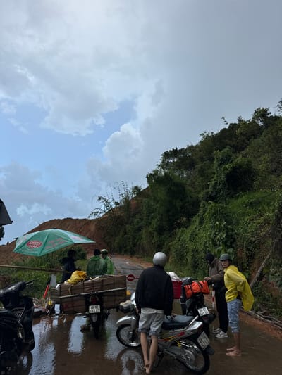 In Nao Sẻ, Vietnam, on a wet, overcast day, a landslide has blocked a road. Several people are gathered near the blockage. In the foreground, three motorbikes are parked on the flooded road. One motorbike is loaded with large cardboard boxes. A green umbrella provides shade for a small stand. The landslide debris, consisting of reddish-brown earth and rocks, covers a significant portion of the hillside. Dense green foliage and trees line the upper right side of the image, contrasting with the bare earth of the landslide. The sky is filled with heavy, gray clouds. The overall atmosphere suggests a disrupted scene due to natural forces.