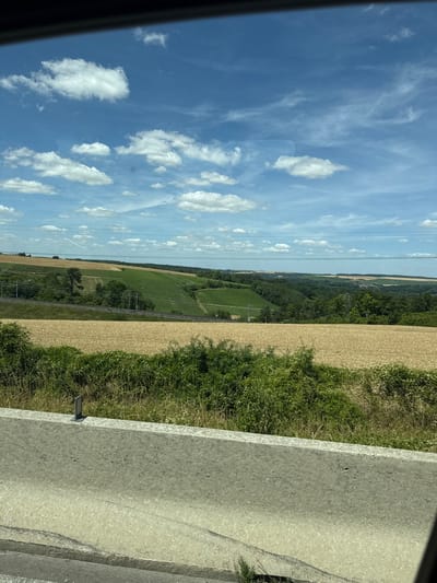 The image captures a landscape scene, likely from a moving vehicle, with a concrete barrier in the foreground. Beyond the barrier, a field of golden wheat stretches across the mid-ground. Further in the distance, rolling hills are covered in lush green vegetation, possibly vineyards or other crops, interspersed with trees. A railway track with overhead power lines runs horizontally through the middle distance, suggesting an active transportation route. The sky is a vibrant blue, dotted with fluffy white clouds. The overall impression is a pastoral, rural setting, possibly in the vicinity of Poilly, France, given the agricultural landscape and the presence of vineyards. There are no people or discernible human activity in the frame.