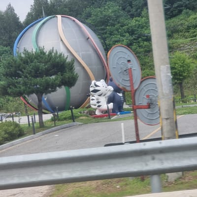 In Bongpyeong-myeon, South Korea, a large, silver, spherical structure with colorful bands wraps around it stands prominently in the background. In front of the sphere, two large mascot statues are visible. The statue on the left is a white tiger with black stripes, in a walking pose. The statue on the right is a dark blue bear-like figure. The mascots appear to be positioned on a red and blue base. To the right of the statues, a metallic pole supports two round, convex mirrors, one above the other, with red framing. The scene is set against a backdrop of lush green trees and a gently sloping hill. A paved road is visible in the foreground, separated from the area with the sphere and statues by a concrete barrier and a metal guardrail. The sky is overcast.