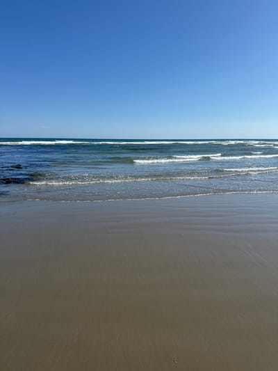 The image depicts a serene beach scene at Ballykinler, United Kingdom. The vast expanse of the clear blue sky dominates the upper portion of the frame. Below, the ocean stretches out, with gentle waves rolling towards the shore. White foam outlines the crests of the waves as they break. The water appears a deep blue in the distance, transitioning to a lighter, more translucent shade closer to the shore. Patches of dark seaweed are visible on the left side of the frame, near the water's edge. The foreground is a wide, flat expanse of wet sand, reflecting the light and showing subtle ripples left by receding waves. The overall impression is one of tranquility and natural beauty.
