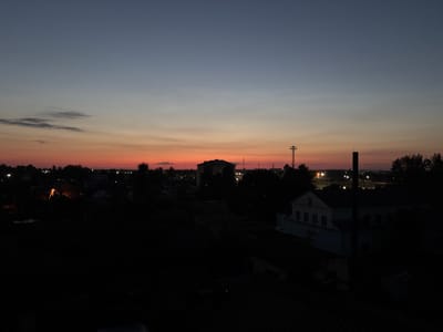 The image captures a dusky, twilight scene over a rural area that is likely part of Пратасевіцкі сельскі Савет, Belarus. The sky exhibits a gradient of colors, transitioning from a deep blue at the top to a warm orange and pink near the horizon, indicating either sunrise or sunset. Silhouetted against this colorful sky are the rooftops of various buildings and the dark shapes of trees. A prominent feature is a radio tower or transmission mast with a structure at its top, located in the mid-ground. Further into the distance, more buildings and lights are visible, suggesting a small settlement or village. The foreground is dominated by the dark silhouettes of rooftops and trees, making it difficult to discern fine details. The overall impression is one of a quiet, rural evening or morning in Belarus.