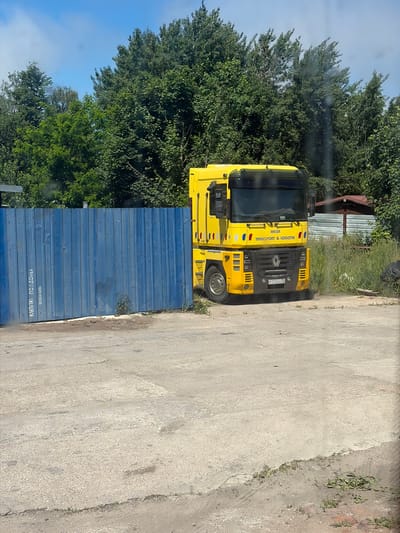 A bright yellow semi-truck is parked on a paved surface next to a blue corrugated metal fence. The truck has "MEISE TRANSPORT & LOGISTIK" written on its side. The license plate is visible and reads "P 778 BM 39". Behind the truck and fence, a dense line of green trees creates a backdrop under a clear blue sky. To the right of the truck, a small wooden structure with a reddish roof is partially visible, along with overgrown grass and vegetation. The ground is rough asphalt with some patches of dirt and gravel.