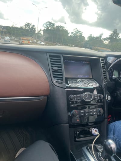 The image provides an interior view of a car, focusing on the dashboard and center console, with a partial glimpse of the outside world. The car's interior features a dashboard with brown, possibly leather, trim on the passenger side and black components for the instrument panel and center stack.

The center console prominently displays an infotainment screen showing "Bluetooth Audio" and the time "16:13". Below the screen, an analog clock is integrated into a panel of control buttons for the audio and climate systems, with visible labels like "MAP", "ROUTE", "MODE", and "DUAL". To the right of the controls, a USB car charger is plugged into an outlet, with a white charging cable extending from it. Partially visible on the right side of the frame are a person's legs, clad in blue jeans and light-colored shoes, suggesting either the driver or a front passenger.

Through the windshield, the exterior scene reveals a bright, cloudy sky dominating the upper left quadrant. Below the sky, a road is visible, flanked by numerous green trees. Several other vehicles, including what appears to be a yellow bus or large truck, are present on the road, along with street lights.

The image captures an activity taking place in a car, possibly while driving or stopped in traffic, with audio playing via Bluetooth and a device charging. The time of day is 4:13 PM, indicated by the screen. The weather is overcast or partially cloudy. The location context indicates this scene is in Theta ward, Kenya. The car's interior design suggests it might be an Infiniti FX or QX70 model.
