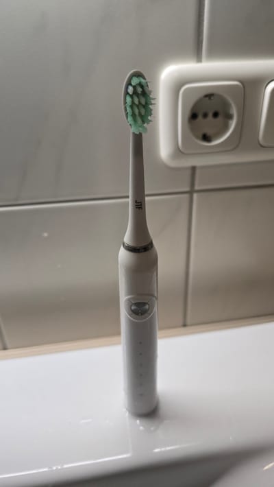 The image displays a white electric toothbrush with a green-bristled brush head, positioned upright on a white sink. The toothbrush has a silver band around its neck and a circular button near the middle of its handle, with several small indicator lights below it. The background consists of a tiled wall and a white electrical outlet with two sockets. The scene appears to be in a bathroom. The image does not provide any information about the city of Lauingen (Donau), Germany, or any people.