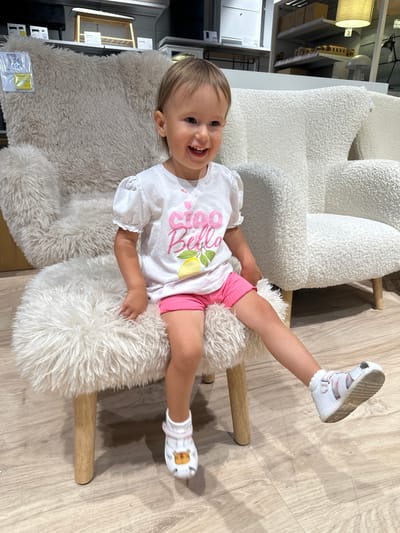 A young child with light brown hair is seated playfully on a white, fluffy stool or pouf in what appears to be a furniture or home goods store in Ustroń, Poland. The child is looking directly at the camera with a wide, joyful smile, showing her teeth. Her right leg is extended upwards, with her white sandal-clad foot visible, while her left leg is bent. She is wearing a white short-sleeved t-shirt with the word "Bella" in pink script and a yellow lemon illustration printed on the front, paired with pink shorts. Her white sandals have pink and white straps and one has a brown character design on the front.
Behind the child is a larger, white chair or sofa with a textured, boucle-like fabric. The floor is made of light-colored wooden planks. In the background, there are metal shelving units displaying various items typical of a home store, including lampshades, boxes, and picture frames. A tag on a separate, lighter beige furry rug on the floor shows the text "SAHARA" and "500 zł", indicating the price in Polish Złoty. A number "2" is also visible on a shelf unit in the upper left background. The overall setting suggests a brightly lit indoor environment during the daytime.