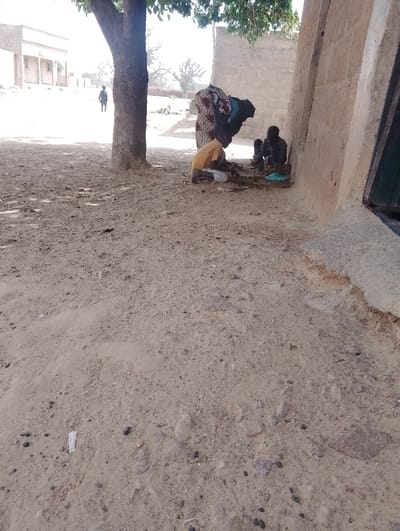 The image depicts an outdoor scene in Kanwa, Nigeria, under bright daylight. The foreground consists of dry, sandy earth with scattered small rocks and debris.
On the right side of the frame, a light-colored, possibly concrete or mud-brick, building wall is visible, extending diagonally upwards from the bottom right corner. A dark, possibly green, door or window frame is partially visible on this wall.
Towards the center-right, under the partial shade of a tree with green foliage, an adult female and two children are situated. The adult female, wearing a multi-patterned dress (red, white, and black) and a dark head covering, is standing and leaning forward, looking down at the ground. To her left and slightly in front, a child wearing a yellow short-sleeved top and light-colored bottoms is crouching or kneeling on the ground, appearing to interact with small objects on the dusty surface. To the adult's right, closer to the building wall, a second child in dark clothing is seated or crouching on the ground. A teal-colored plastic object is near this child.
In the mid-ground, a tree trunk rises centrally, providing shade for the group. Beyond the tree, the ground is open and dusty. In the far background, a light-colored building with multiple openings or windows is visible. To the left of this building, a solitary individual in dark clothing is walking away from the camera. Sparse, dry vegetation and distant trees are visible under a bright sky. The overall environment suggests an arid or semi-arid climate.