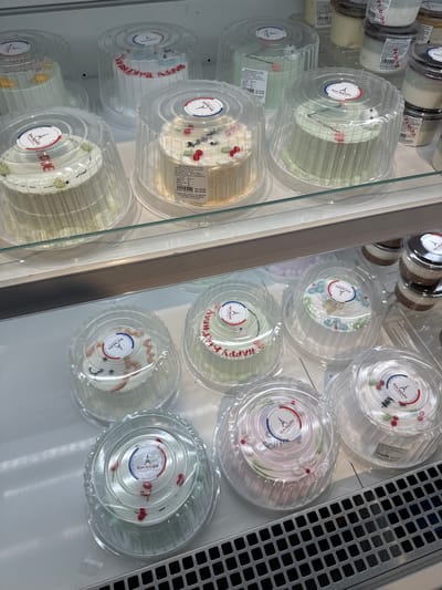 A retail display case contains multiple small, round cakes in clear plastic containers. The cakes are decorated with frosting and some have fresh fruit or edible flowers. Labels with a French-themed logo, "Bon Appétit," are affixed to the top of each container. The display case is lit from above, illuminating the cakes and creating reflections on the plastic packaging. The background is a clean, white surface, typical of a bakery or dessert shop. The overall impression is one of freshness and visual appeal. It is likely a bakery or dessert shop in Phu Thuy, Vietnam, given the context provided.