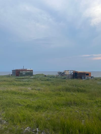The image depicts an outdoor scene with two dilapidated structures situated on a grassy incline overlooking a body of water. The sky is a pale blue with scattered white clouds, suggesting it is daytime, likely late afternoon or early evening given the soft lighting. The weather appears to be calm and possibly overcast, as there are no strong shadows.
The first structure, closer to the viewer and towards the bottom of the frame, is a rectangular building with a green, rusty roof and metallic siding that appears to be partially weathered and damaged. It has a pipe extending from its roof. Adjacent to it is a smaller, darker structure.
Further up the incline and to the right, another structure is visible. This one is larger and appears to be made of metal, with an orange-brown exterior. It is also in a state of disrepair, with broken windows and visible structural damage, including remnants of scaffolding or support beams. Debris is scattered around this structure.
The ground is covered in lush green grass, with some patches of lighter vegetation and small white wildflowers. The terrain slopes downwards towards a calm, grey body of water that stretches to the horizon. The overall impression is one of abandonment and a remote, perhaps industrial, setting. The image evokes a sense of solitude and the passage of time, with the decaying structures standing as remnants of past activity in a natural environment. There is no visible text in the image. The location context suggests Yamburg, Russia, which aligns with the rugged, open landscape and the presence of industrial-style structures.