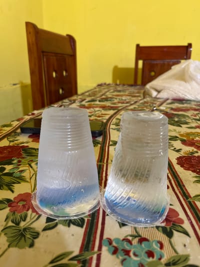 Two clear plastic cups filled with a blue and green liquid are placed on a floral-patterned tablecloth. The cups appear to be partially frozen, with ice forming on the exterior. In the background, two wooden chairs with a yellow wall behind them are visible. The setting is indoors, likely a dining area, and the overall scene suggests a casual, home environment. The image does not provide enough information to determine the specific location within Tanjung Pura, Indonesia, or the actions or interactions of any individuals.