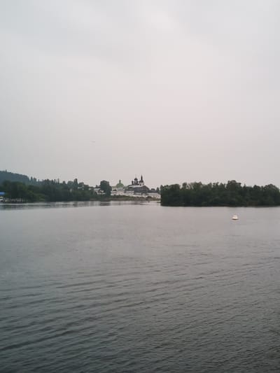 An overcast, grey sky dominates the upper portion of the image, with a faint hazy horizon. In the middle ground, a body of water, likely a river, stretches horizontally across the frame. The water's surface is rippled, suggesting a gentle current. Along the far bank of the water, a treed shoreline is visible, dotted with buildings. On the left, a cluster of buildings with white walls and dark roofs appears to be part of a settlement, possibly with a blue-roofed structure. Further to the right, a more prominent complex of buildings with domes and spires is visible, indicative of a religious or historical site. A small island densely covered with green trees is situated in the water on the right side of the frame, and a white and orange buoy floats near it. The overall scene suggests a serene, perhaps slightly melancholic, landscape on a cloudy day. The location is the city of Goritsy, Russia, likely showing the Goritsky Monastery.
