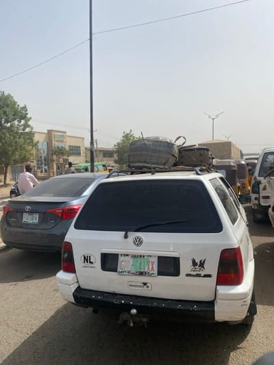 The scene depicts a congested street in Gobirawa, Nigeria, under a hazy daylight sky. In the foreground, a white Volkswagen Golf station wagon is observed from the rear, displaying a Jigawa state license plate "GH 404 XX". The vehicle has dark-tinted rear windows, "NL" and "Joker" stickers to the left of the license plate, and a "SportX" decal with a winged creature on the right. A prominent roof rack carries a striped fabric bag and a green hard-shell case, among other indistinct items. A trailer hitch is visible below the rear bumper. Directly behind the white Volkswagen is a dark grey Toyota Corolla sedan, bearing a Jigawa state license plate "FGE 18CW". A person in a light-colored shirt is seated on a motorcycle positioned behind the Toyota Corolla, facing away from the camera. To the right, a queue of vehicles includes multiple yellow and black commercial tricycles. On the left side of the frame, a green small bus or van is partially visible. In the background on the left, a multi-story building with "CRONIL Hotel" visible on its facade stands amidst sparse trees. A tall, dark utility pole occupies the mid-ground, with power lines traversing the upper portion of the frame. Distinctive streetlights with horizontal cross-arms are visible in the distant background on the right.