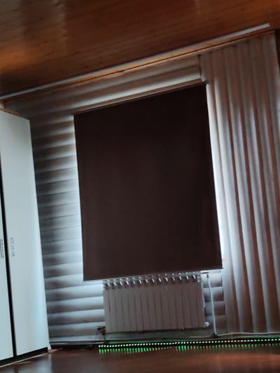 The image depicts an indoor scene with a wooden paneled ceiling and walls. A large, dark brown roller blind is lowered over a window, obscuring the view outside. To the right of the blind, a light-colored, pleated curtain is visible. Below the window, a white radiator is installed. In the lower portion of the image, a dark wooden floor reflects the light from a strip of green LED lights. To the left of the scene, a portion of a white cabinet or wardrobe is visible, featuring two silver handles. The lighting suggests an interior space, possibly a room or hallway. The specific location in Solnechnyi, Russia, cannot be determined from the visual information provided in the image.