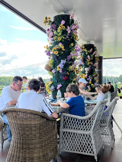 A group of people are seated at tables on an outdoor patio adorned with large, ornate floral columns. The patio is covered by a white roof structure, and in the background, a cloudy sky, trees, and distant structures are visible.

In the foreground, a man wearing a white t-shirt and glasses is seated at a table, engaged in conversation with a young person in a white shirt with black stripes. To their right, a boy in a blue t-shirt is seated at another table, with a woman in a white cap and tank top attending to a child with red hair.

The tables are covered with white tablecloths and set with plates and a bottle of water. The chairs are made of woven wicker. The floral columns are substantial, covered in dense greenery and decorated with a vibrant assortment of artificial flowers in various colors including purple, yellow, pink, and white. Some small wicker baskets are also attached to the columns.

The scene appears to be a leisurely gathering or meal at a restaurant or cafe. The overall atmosphere is relaxed and summery. The location is identified as Akhlebaikha (Kovriginskii S/S), Russia.