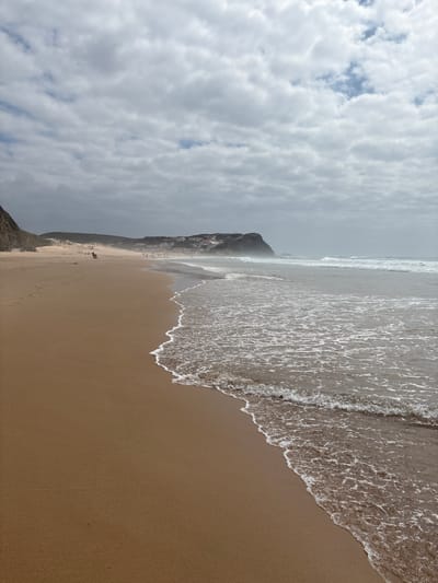 This is a vertical, eye-level shot of a wide, sandy beach with the ocean on the right. In the foreground, a gentle wave breaks on the wet sand. The sand is a light brown color and appears smooth, with some footprints visible in the distance. The ocean is a muted gray-blue and shows small, choppy waves with white foam.

In the middle ground, the beach stretches out towards a distant coastline. Several people are scattered across the sand, some walking and others sitting. A small group of buildings with white walls and red-tiled roofs is visible on the hills in the background. A rocky outcrop juts out into the sea on the far left of the image.

The sky is overcast with heavy, gray clouds that partially obscure the sun, giving the scene a muted, diffused light. The overall atmosphere is calm and somewhat subdued, characteristic of a cloudy day at the beach. The location is likely the Praia da Arrifana in Aljezur, Portugal, a popular surf spot known for its dramatic cliffs and golden sands.
