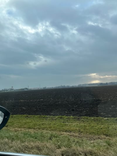 The image depicts a rural landscape in La Wantzenau, France, under an overcast sky. In the immediate foreground, the lower-left corner is partially obscured by the black frame of a vehicle's side mirror. Below the horizon, a strip of green and dry brown grass runs horizontally across the lower portion of the frame. Beyond this, a vast expanse of dark brown, tilled agricultural field dominates the midground, showing visible furrows and disturbed soil. On the distant horizon, a line of structures is visible, including a church spire with a white building to its left, multiple commercial or residential buildings, and several utility poles or towers. A highway or major road runs horizontally across the background, featuring several vehicles, including a large white truck moving from left to right. The sky is largely covered with grey clouds, but a prominent patch of sunlight breaks through on the far right, illuminating a section of the horizon and casting a brighter glow on some distant elements. No discernible individuals or specific interactions are present.
