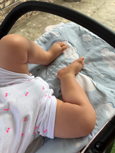 A close-up, high-angle shot shows the legs and feet of a baby lying on a light blue blanket with a cloud and star pattern. The baby is wearing a white onesie with small pink cherry motifs. To the left of the baby's legs, a black handle of a stroller is visible. The background appears to be an outdoor setting, possibly a patio or sidewalk, with a textured surface and a hint of greenery. The image does not contain enough information to determine the specific location within Rusokastro, Bulgaria.