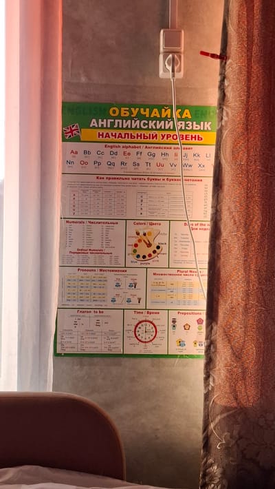 The image shows a wall with a brightly colored educational poster in Russian titled "ENGLISH EDUCATIONAL LANGUAGE. ELEMENTARY LEVEL." The poster features the English alphabet with phonetic transcriptions, sections on numbers, colors, pronouns, verbs, time, and prepositions, along with illustrations. To the right of the poster, a brown patterned curtain is visible. The poster is hanging on a light gray wall, and there is a white electrical outlet with a plug in the wall to the right of the poster, suggesting an indoor setting. The lighting appears to be natural, possibly from a window, creating soft shadows. The presence of the poster and curtain indicates a domestic or educational environment. The location of Novoilinsky, Russia is not discernible from the visual information in the image itself.