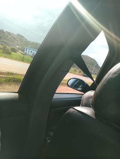 The image, taken from inside a car, shows a road and a hilly landscape. A billboard with images of three men is visible on the left side of the road. The men appear to be politicians, with text in English and what seems to be an African language. The billboard is accompanied by text that reads "COMPLETING PROGRESS. WE CANING SHE PERMANEN. RUFUS". The scene appears to be set in a rural or semi-rural area, with vegetation and rocky hills in the background. The sky is overcast, suggesting it might be daytime with cloudy weather. The interior of the car, with a seatbelt and leather seats, indicates that someone is either driving or a passenger. The overall impression is that of a journey through a Nigerian landscape, possibly during an election campaign period.