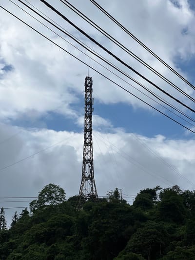The image shows a tall, lattice telecommunications tower situated on a densely forested hillside. The tower is dark and metallic, with visible antennas and equipment. Several thick, dark power lines stretch diagonally across the upper portion of the frame, obscuring parts of the sky. The sky is partly cloudy, with patches of blue visible amongst the white and gray clouds. The hillside is covered in lush green trees and foliage, with the tower appearing to be built into the natural landscape. The scene is set in Quản Bạ Commune, Vietnam.