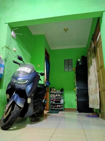 A blue Yamaha motorcycle dominates the foreground of a room with green walls and white tiled floor. Behind the motorcycle, there is a small, four-tier shoe rack filled with shoes. A small, square window can be seen on the green wall. To the right of the window, there's a dark wardrobe and a doorway with a light-colored curtain. Above, a white ceiling with a decorative light fixture is visible. A white WiFi router is fixed to the green wall to the left of the motorbike.