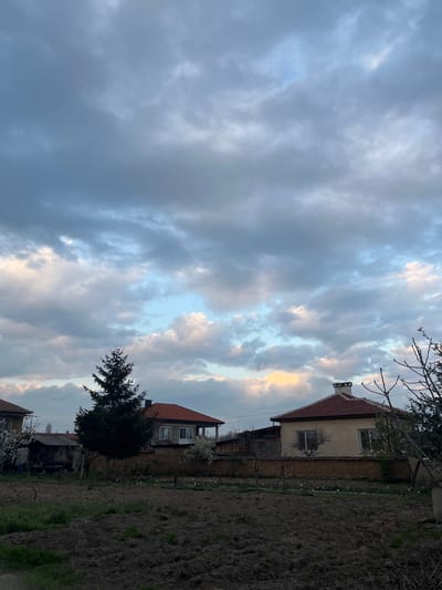 The image depicts a landscape in Panicheri, Bulgaria, under a cloudy sky with patches of sunlight breaking through. A multi-story house with a reddish-brown tiled roof is visible on the right, adjacent to what appears to be a tilled garden. A small tree stands near the base of the house. The image is rotated, with the sky filling the left portion and the ground on the right.