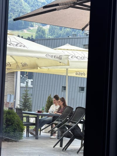 The image shows an outdoor seating area of a cafe or restaurant, possibly in a mountainous region given the background. Two young women are seated at a table. One woman, wearing a dark patterned top and grey pants, is leaning forward, looking down at the table. The other woman, in a white top, is also looking down, and her face is partially obscured. The table has bottles of drinks and other items on it.
Several large, light-colored umbrellas with the Coca-Cola logo are set up, providing shade. In the background, there is a modern building with grey siding. Beyond the building, a lush green, tree-covered hillside is visible, suggesting a natural, possibly rural or semi-rural, location. The overall impression is of a casual daytime scene.
The image appears to be taken through a window or opening, as indicated by the dark framing at the top and bottom of the picture. The lighting suggests it is daytime, likely sunny. There is no explicit indication of emotion, but the setting and activity suggest a relaxed atmosphere. The visible text includes "Coca-Cola" on the umbrellas. The location context provided is Trnovo Municipality (FBiH), Bosnia and Herzegovina, which aligns with the mountainous scenery.